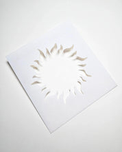 White square plastic pastry stencil featuring a cutout sun shape with wavy, radiant rays.