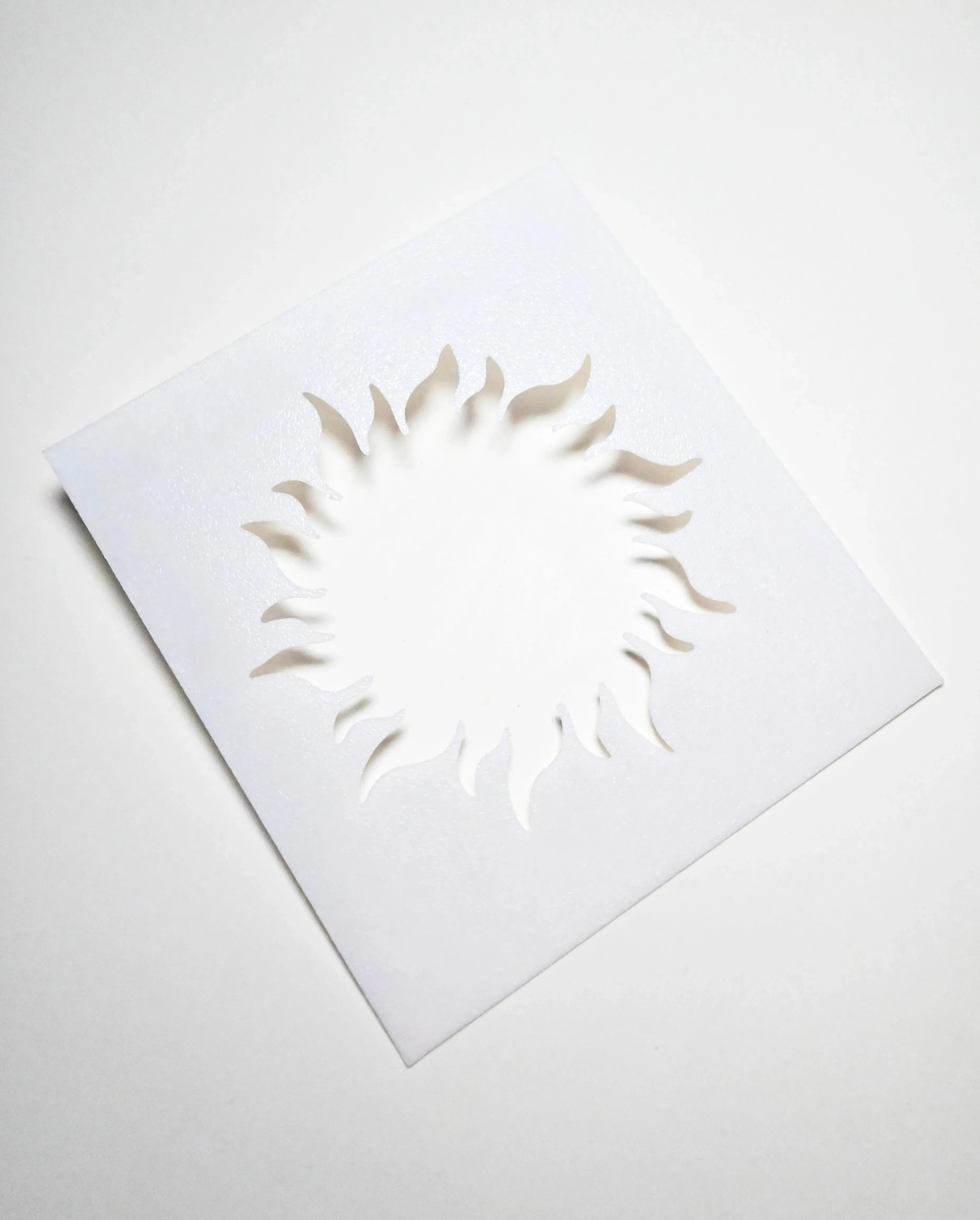 White square plastic pastry stencil featuring a cutout sun shape with wavy, radiant rays.