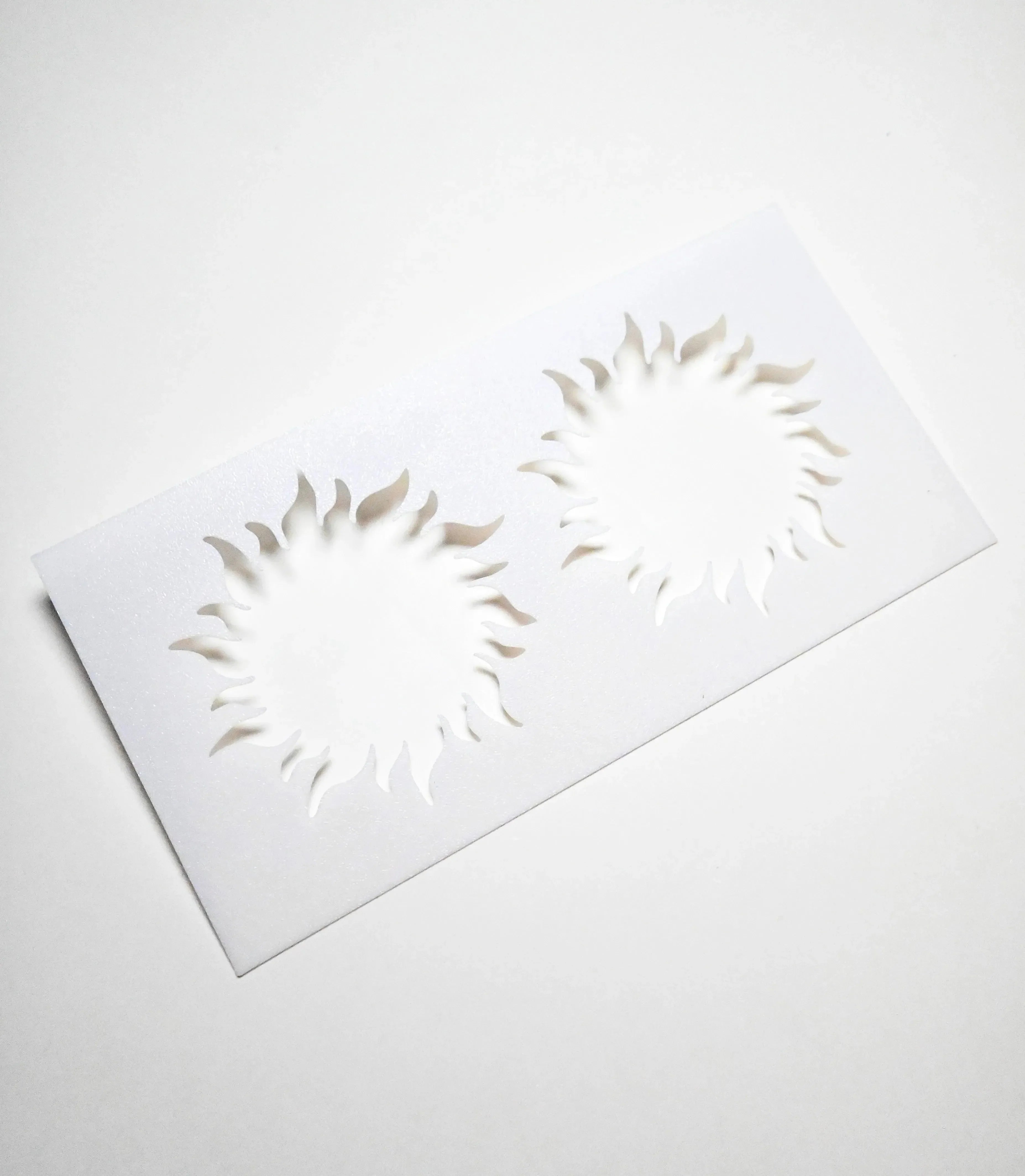 White plastic pastry stencil featuring two detailed sun-shaped cutouts with wavy rays.
