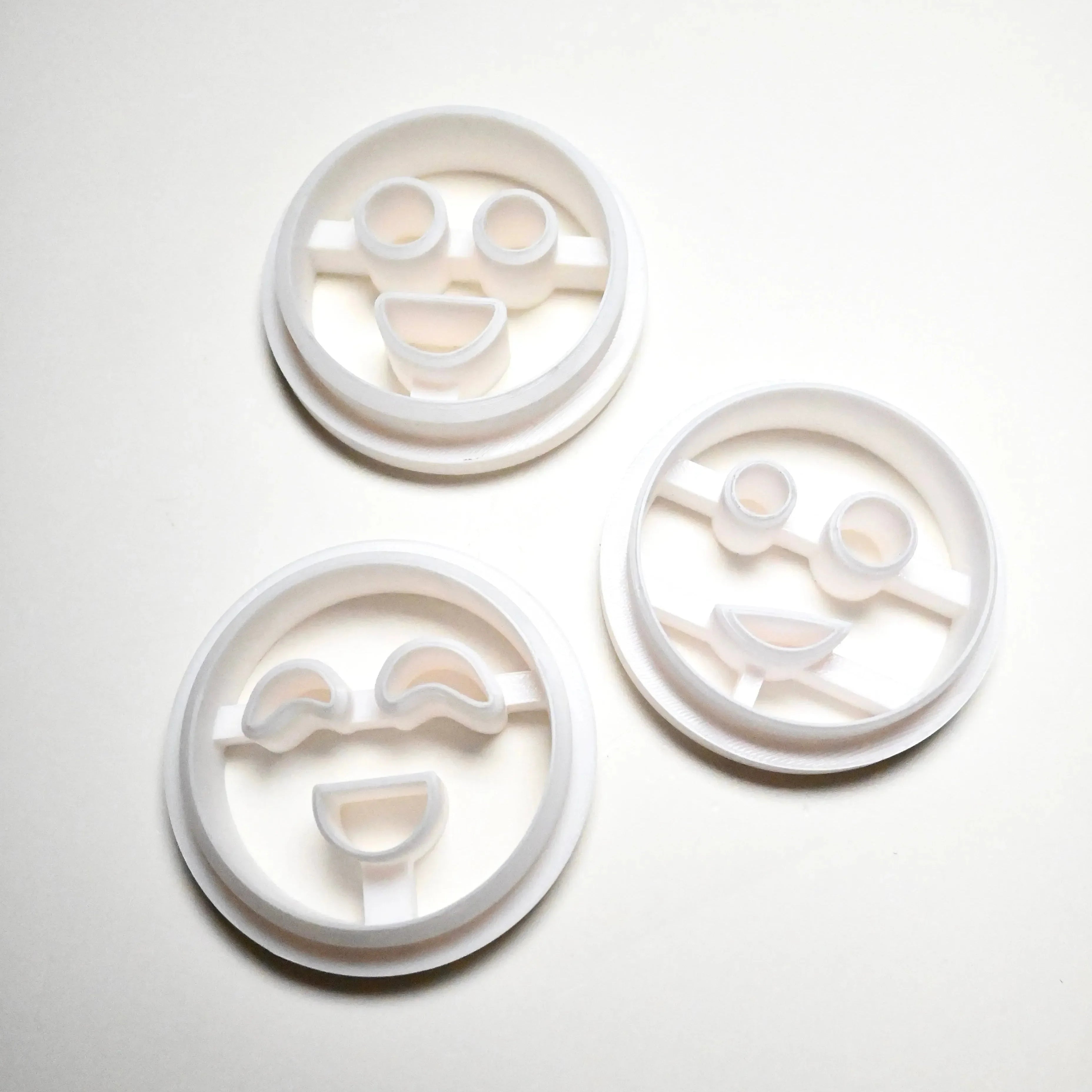 Three white plastic round cookie cutters with different smiley face designs.