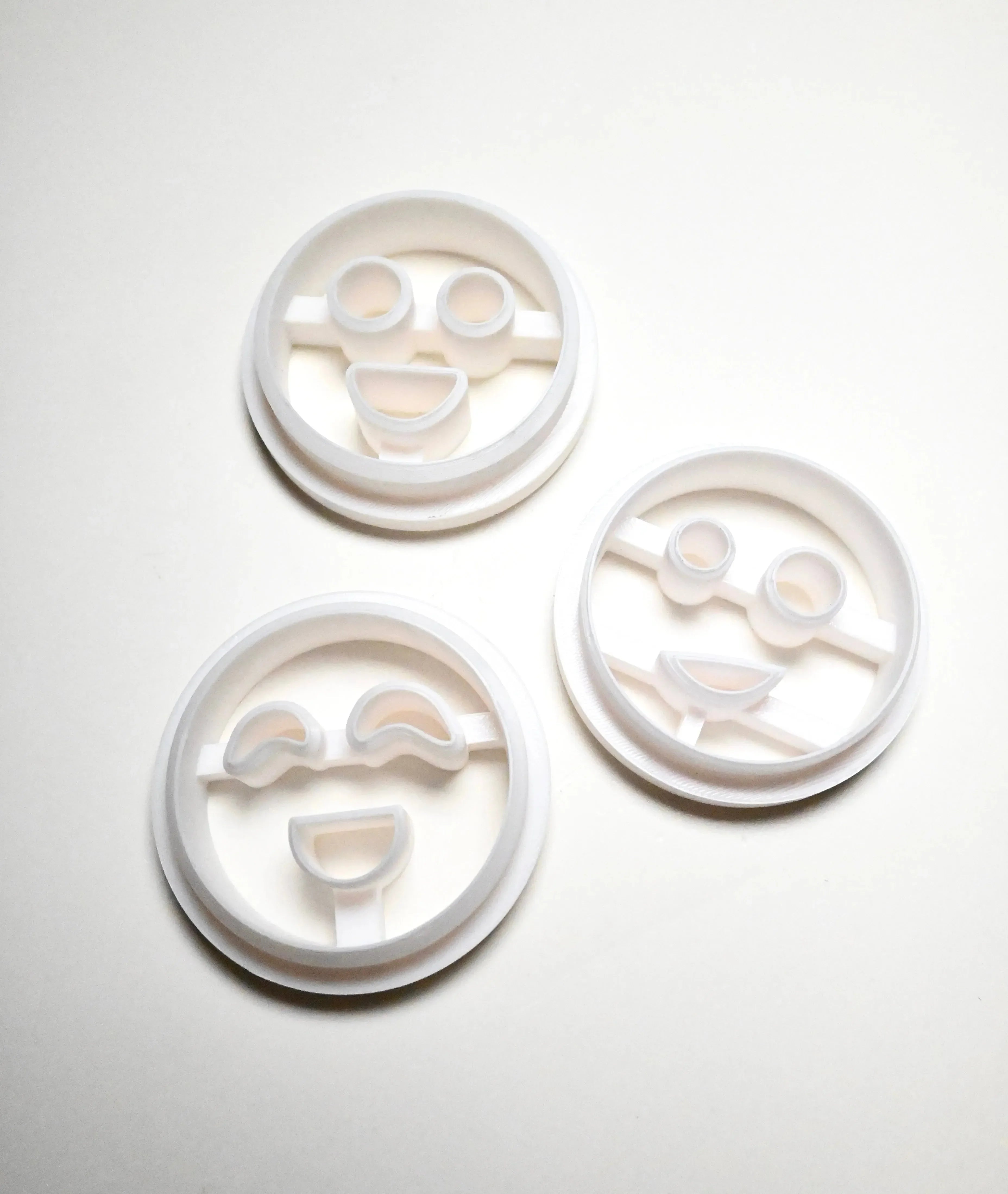 Three white plastic round cookie cutters with different smiley face designs.