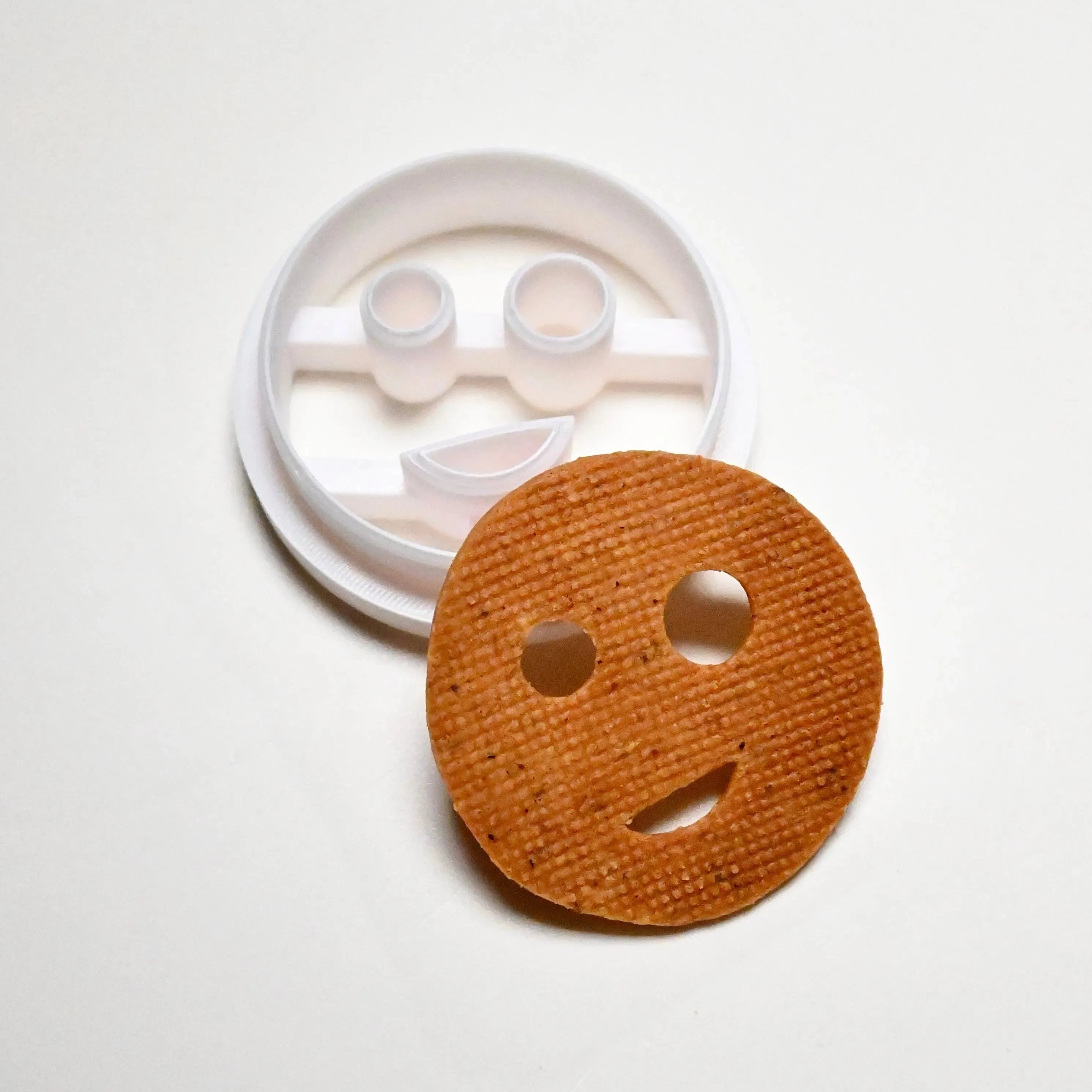 White plastic smiley face cookie cutter next to a round, baked smiley face cookie.