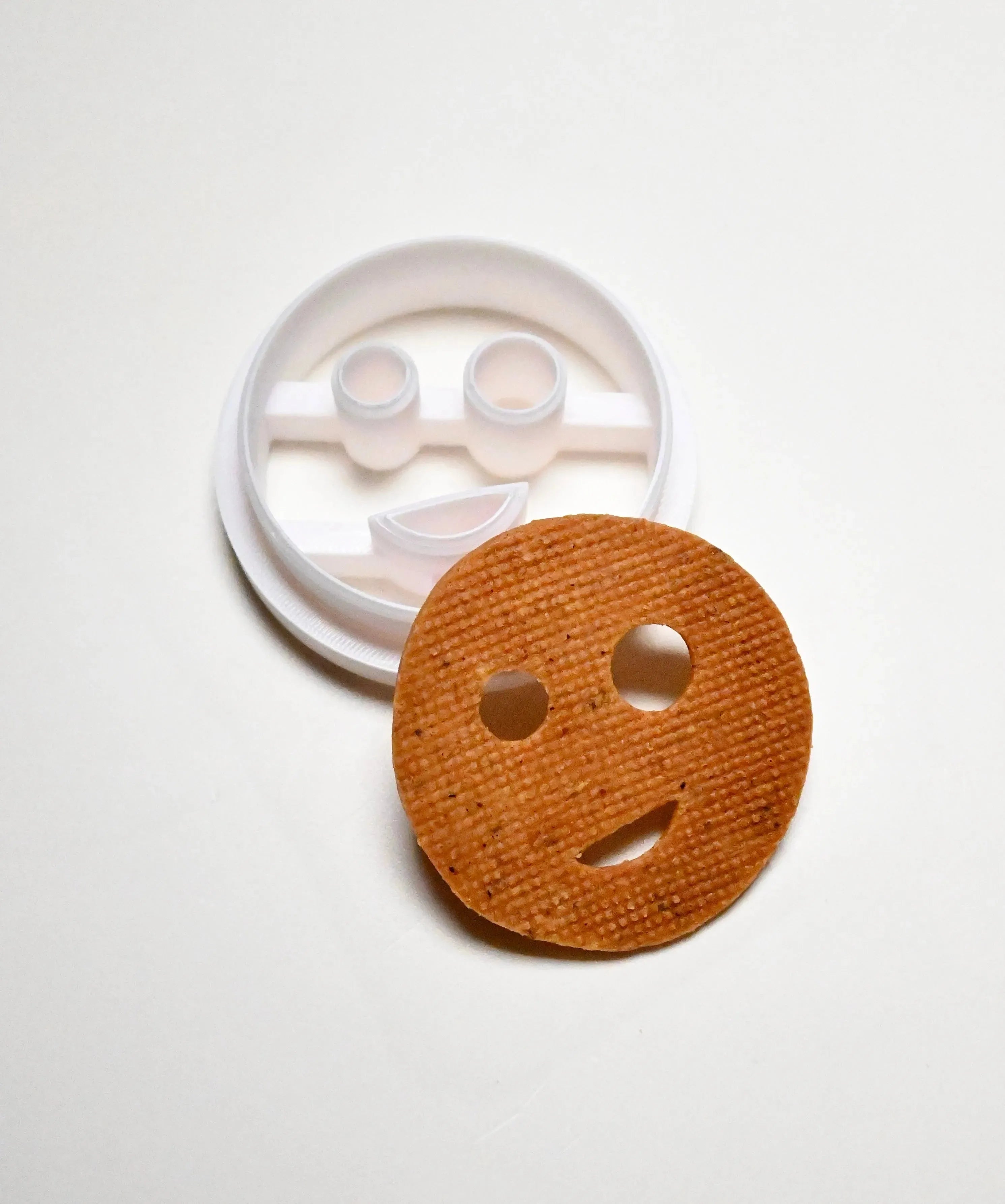 White plastic smiley face cookie cutter next to a round, baked smiley face cookie.
