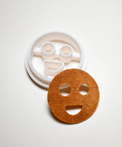 White plastic round smiley face cookie cutter next to a baked circular smiley face cookie.