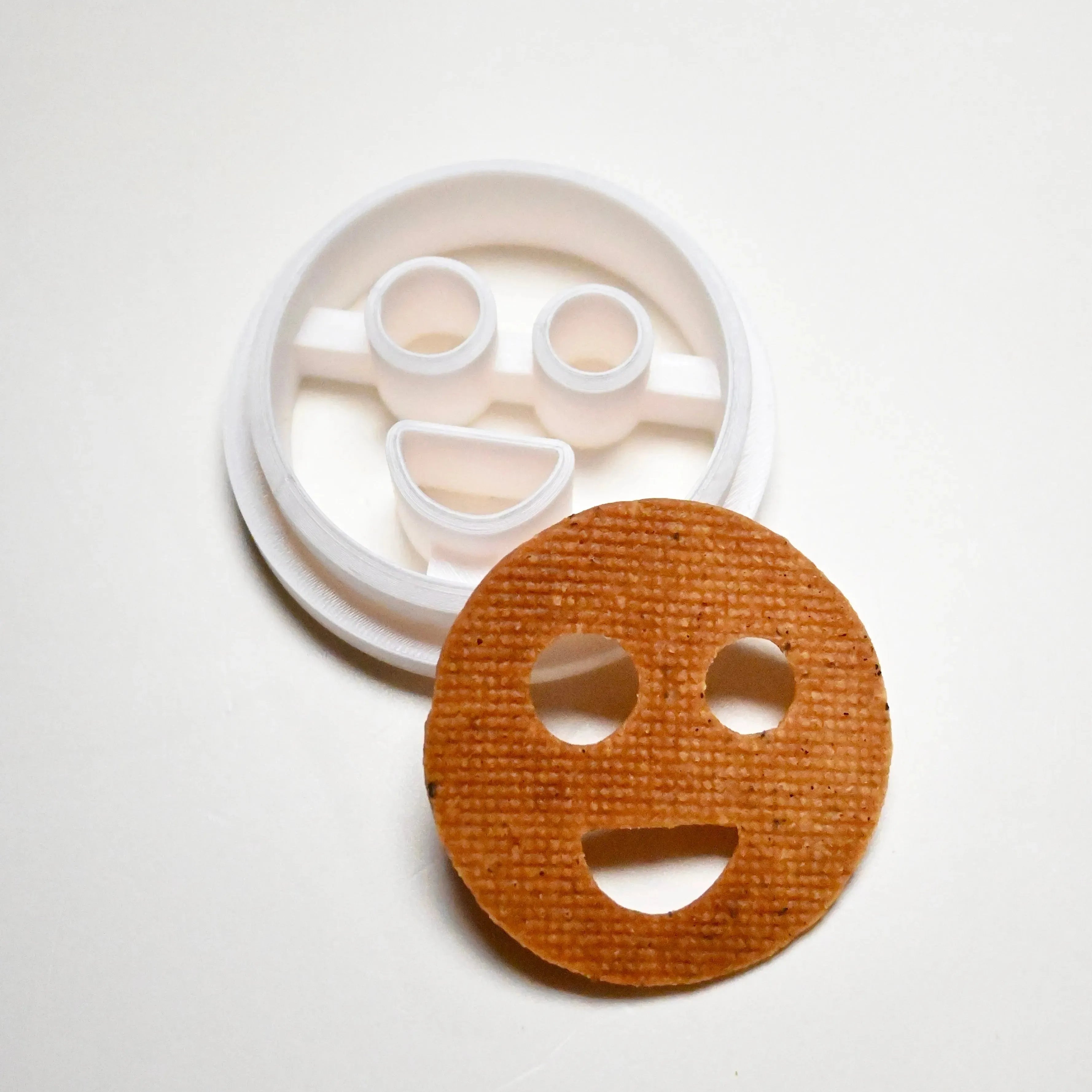White plastic round smiley face cookie cutter next to a baked circular smiley face cookie.