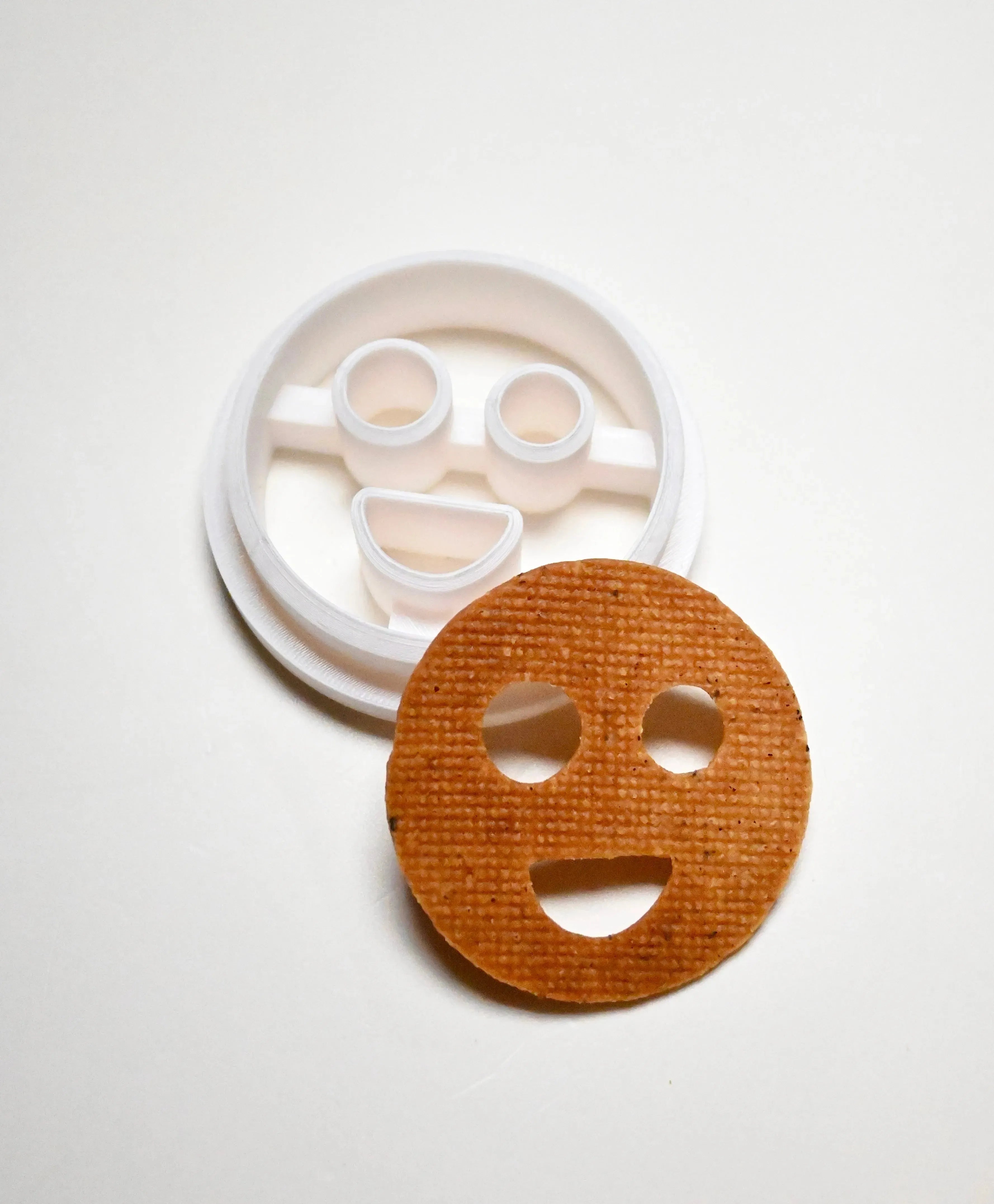 White plastic round smiley face cookie cutter next to a baked circular smiley face cookie.
