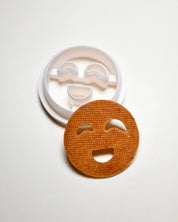 White plastic smiley face cookie cutter next to a round, baked cookie with cut-out happy eyes and mouth.