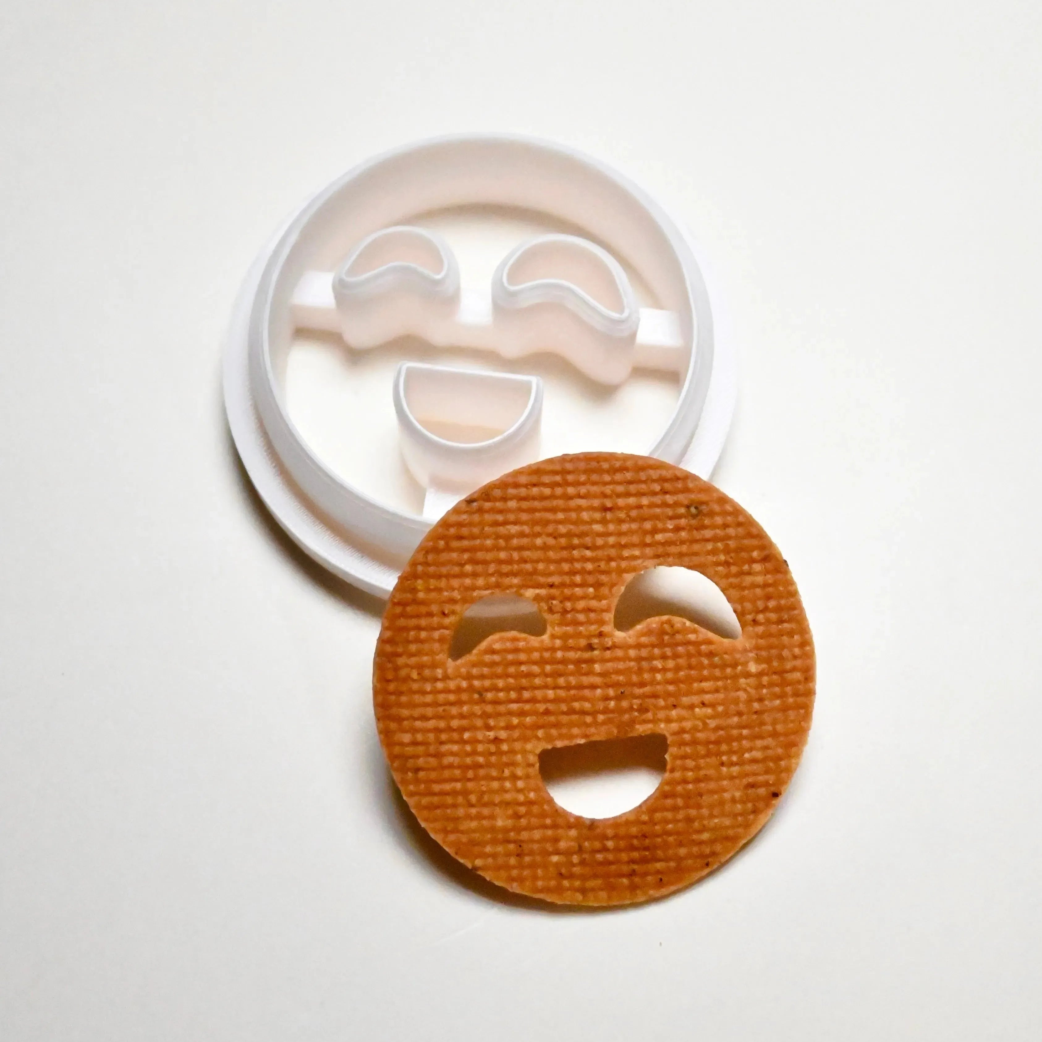 White plastic smiley face cookie cutter next to a round, baked cookie with cut-out happy eyes and mouth.