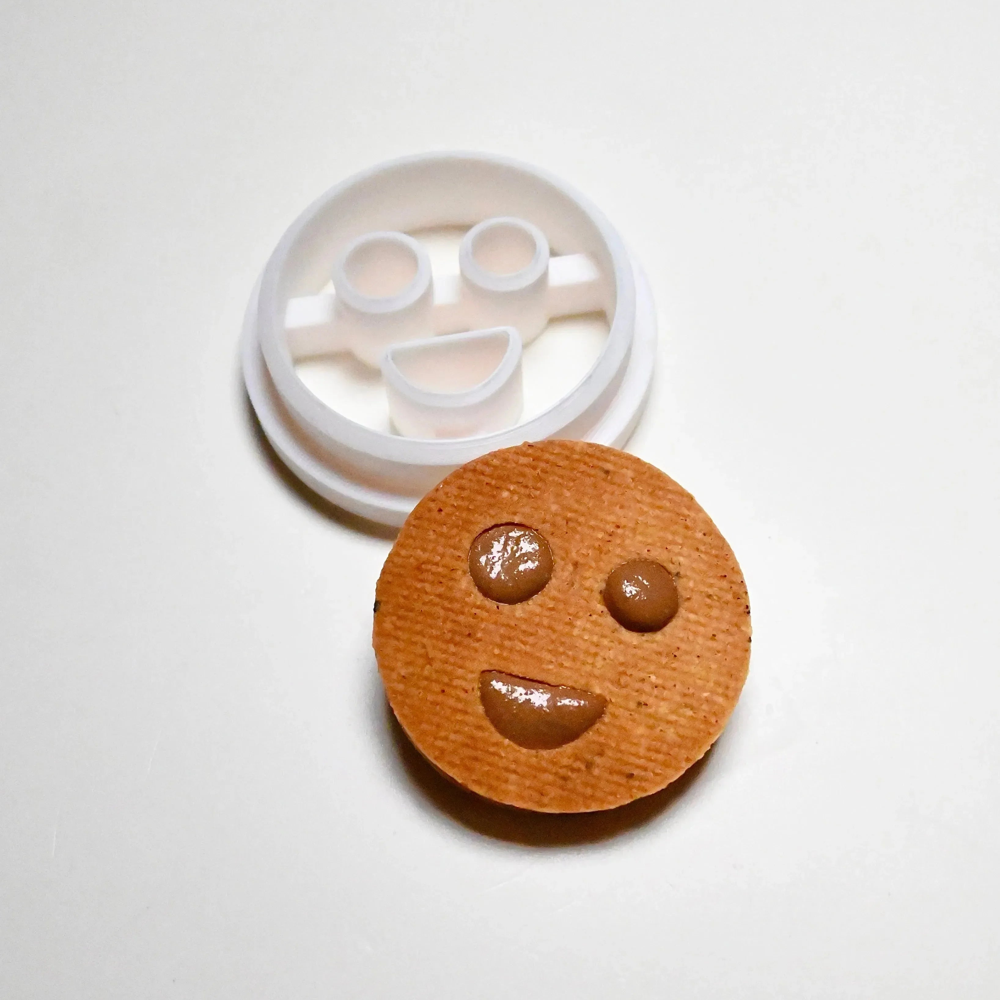 White plastic round smiley face cookie cutter next to a baked cookie with a matching happy face design.
