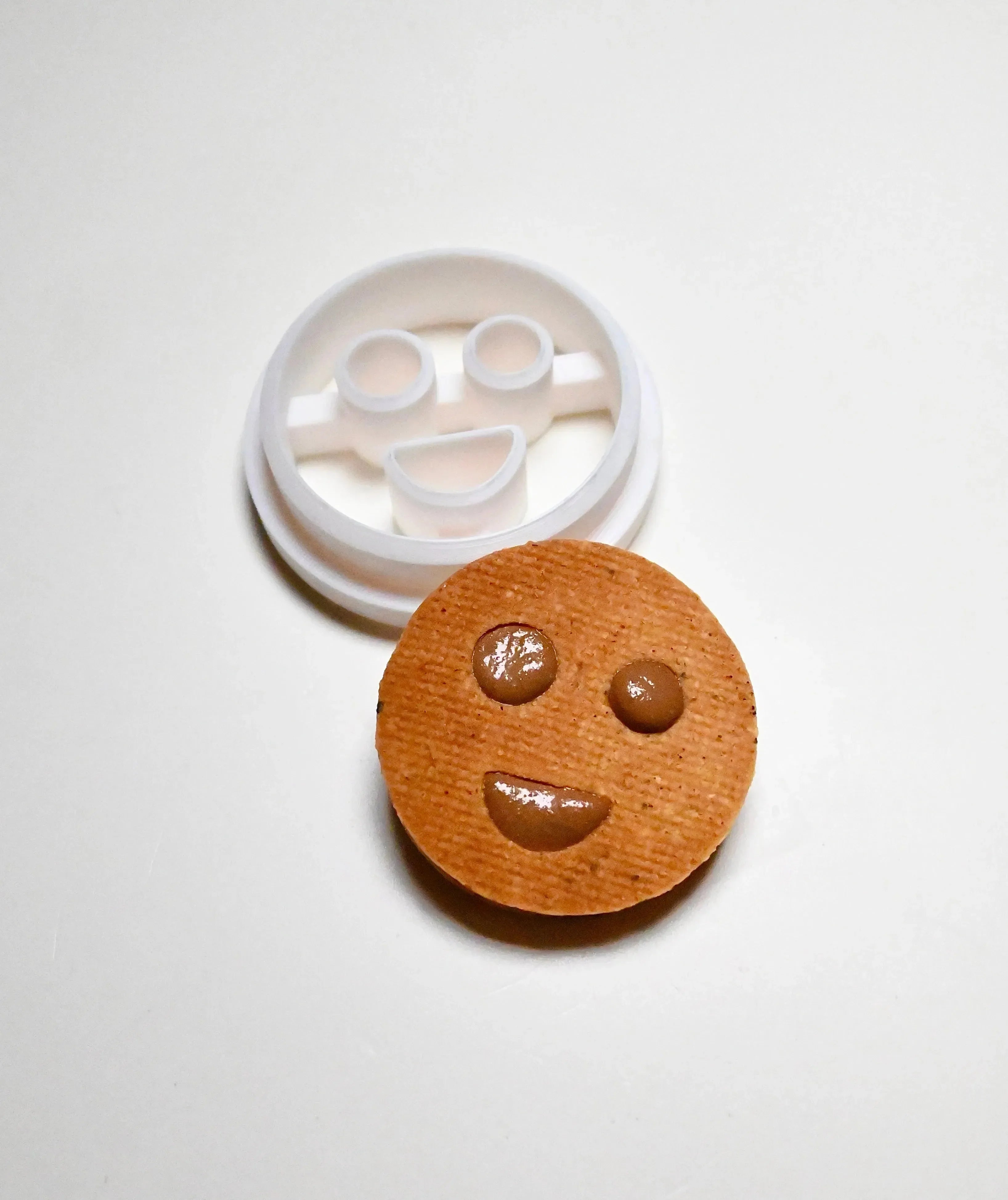 White plastic round smiley face cookie cutter next to a baked cookie with a matching happy face design.