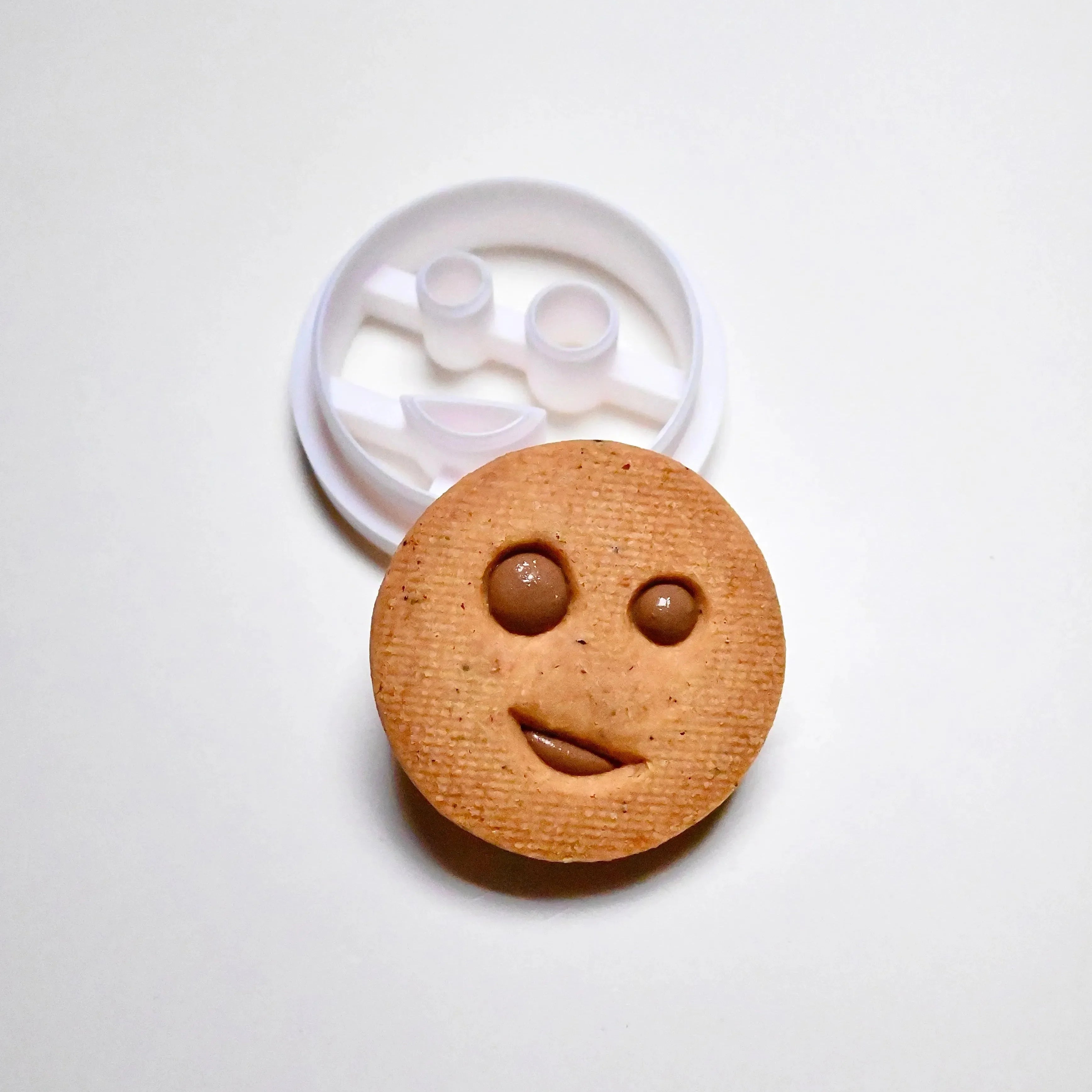 White plastic round smiley face cookie cutter next to a baked round cookie with a winking face.
