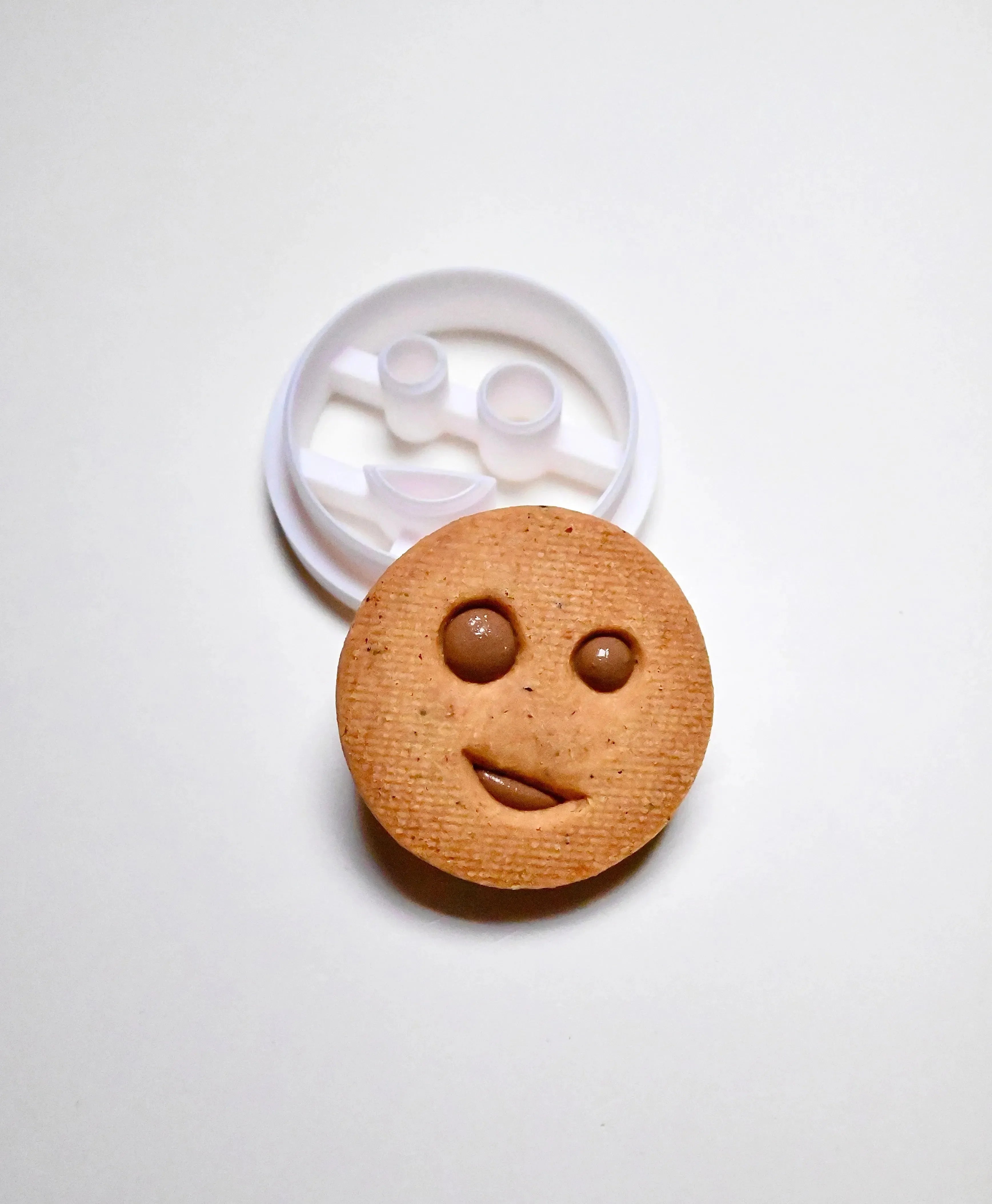 White plastic round smiley face cookie cutter next to a baked round cookie with a winking face.