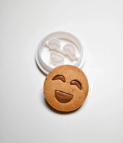White plastic smiley face cookie cutter next to a round, happy face cookie with a chocolate filling.