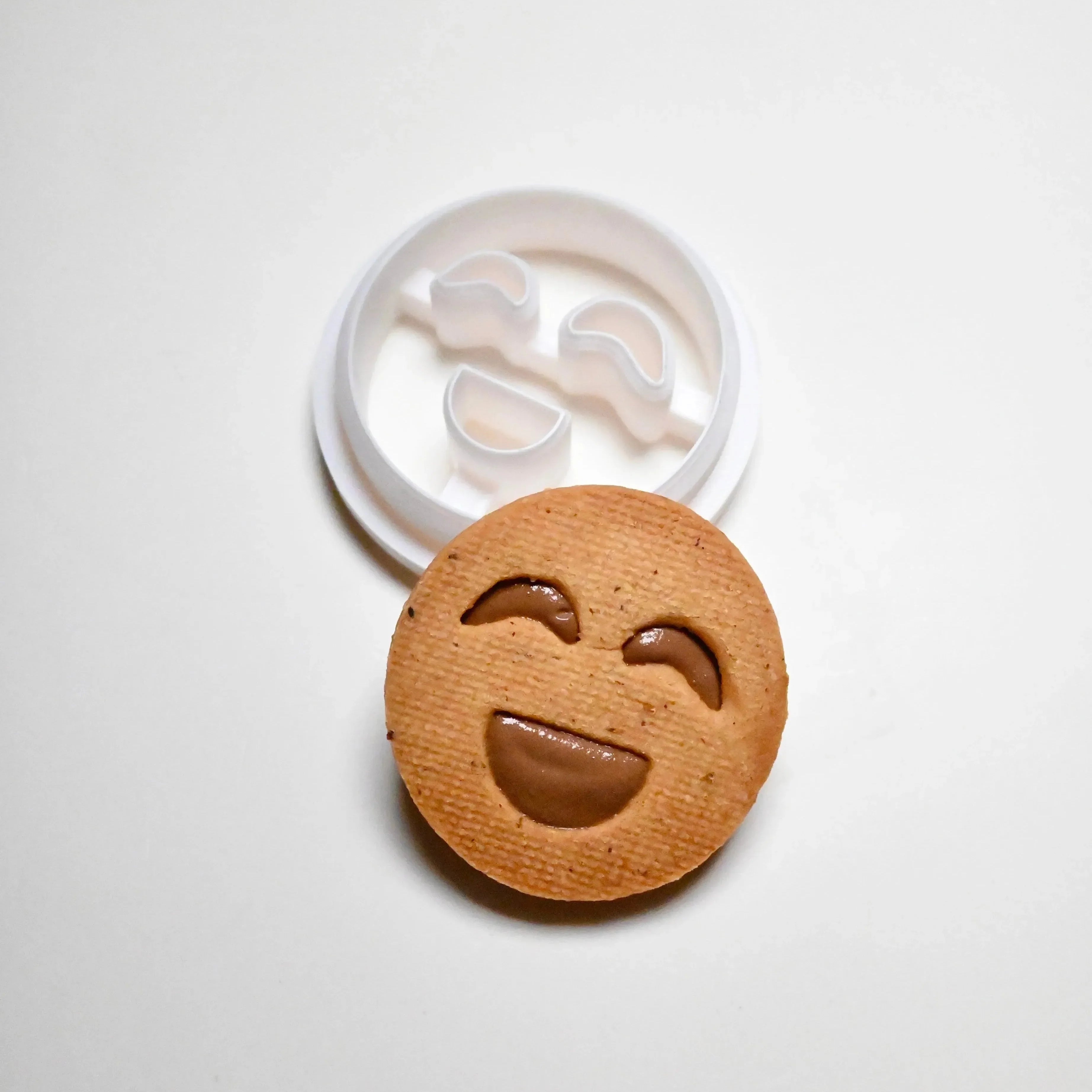White plastic smiley face cookie cutter next to a round, happy face cookie with a chocolate filling.