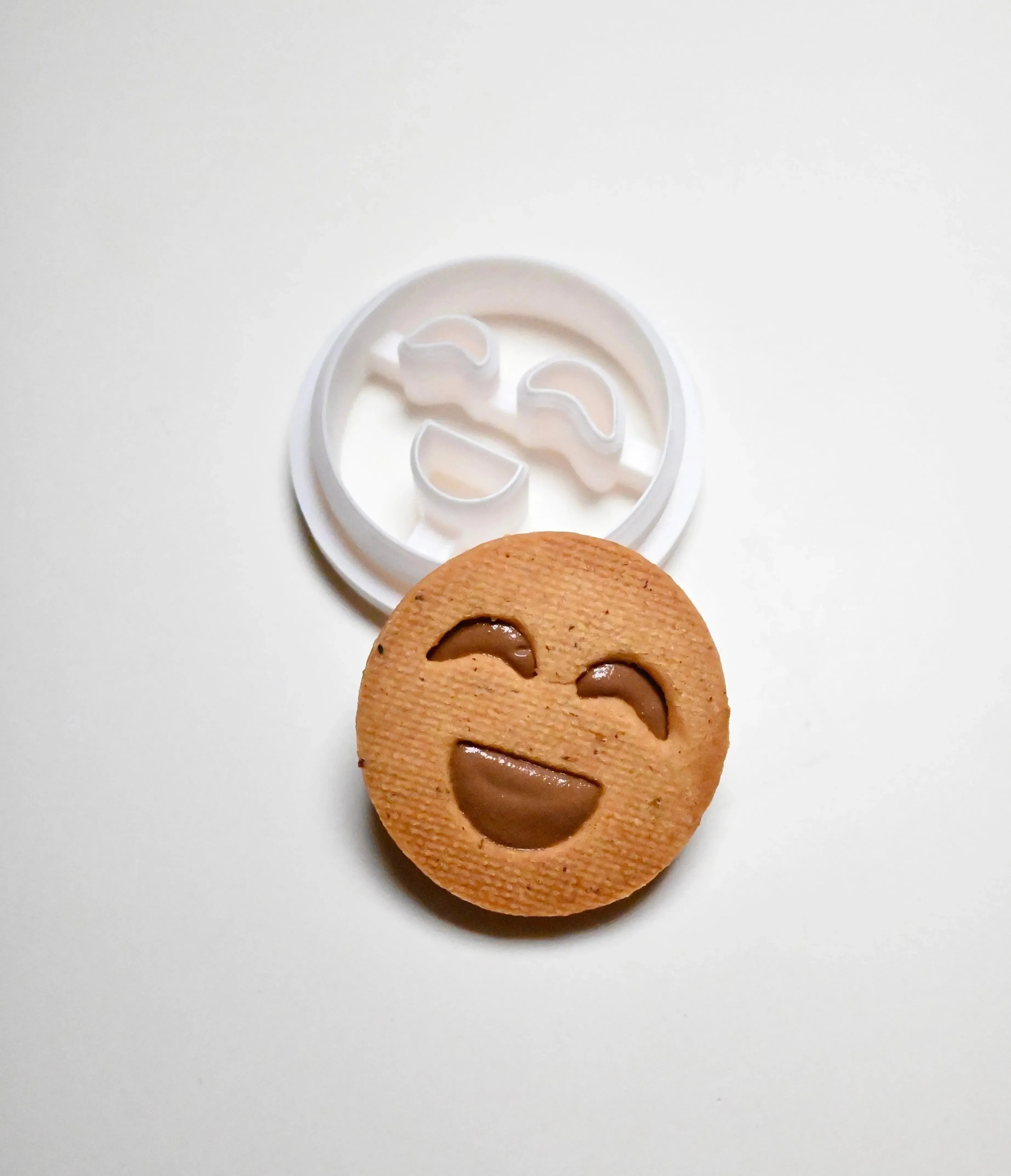 White plastic smiley face cookie cutter next to a round, happy face cookie with a chocolate filling.
