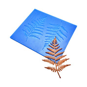 Blue silicone mat with two fern leaf molds next to a brown, leaf-shaped tuile.
