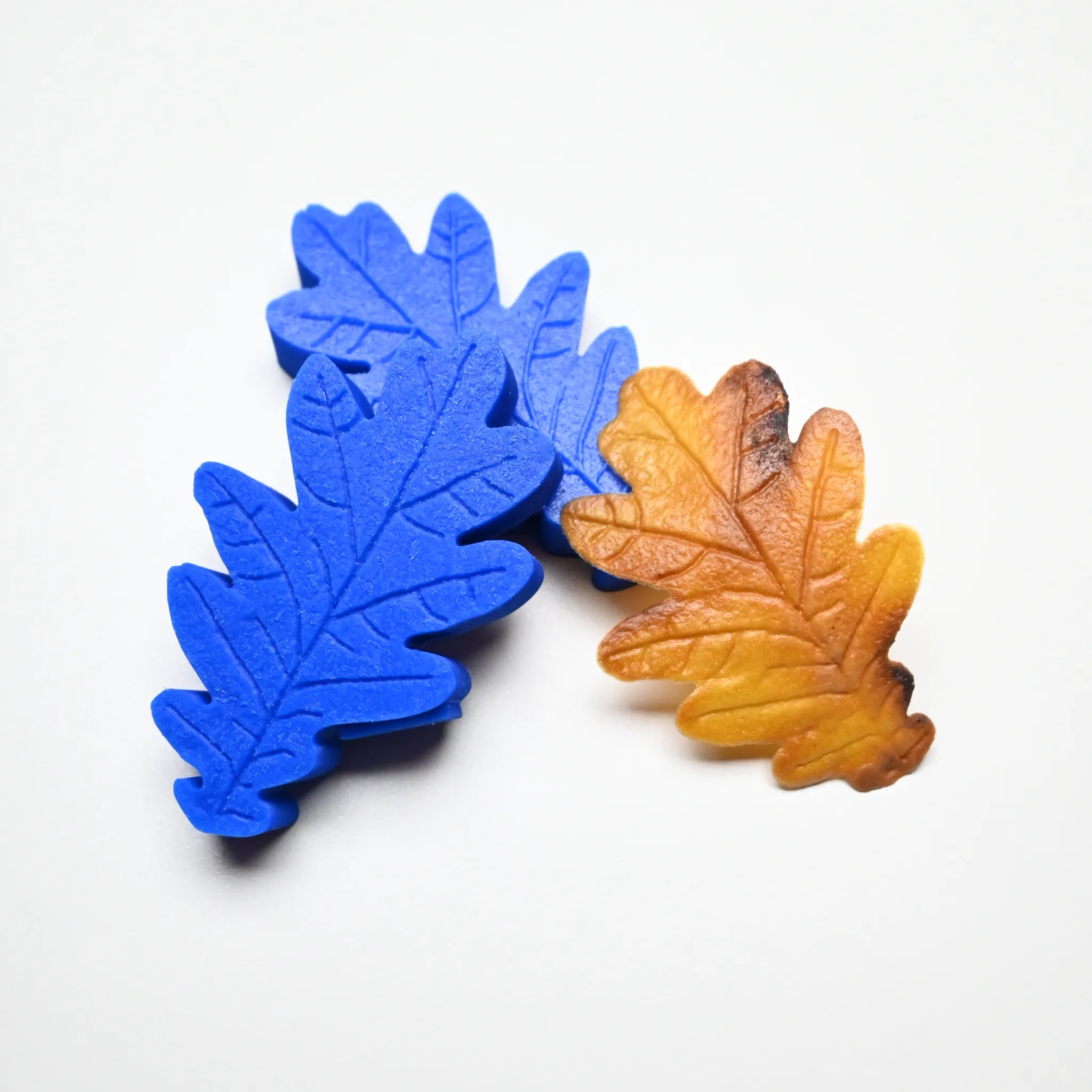 Two blue silicone oak leaf molds with detailed veins next to a realistic brown pastry leaf.